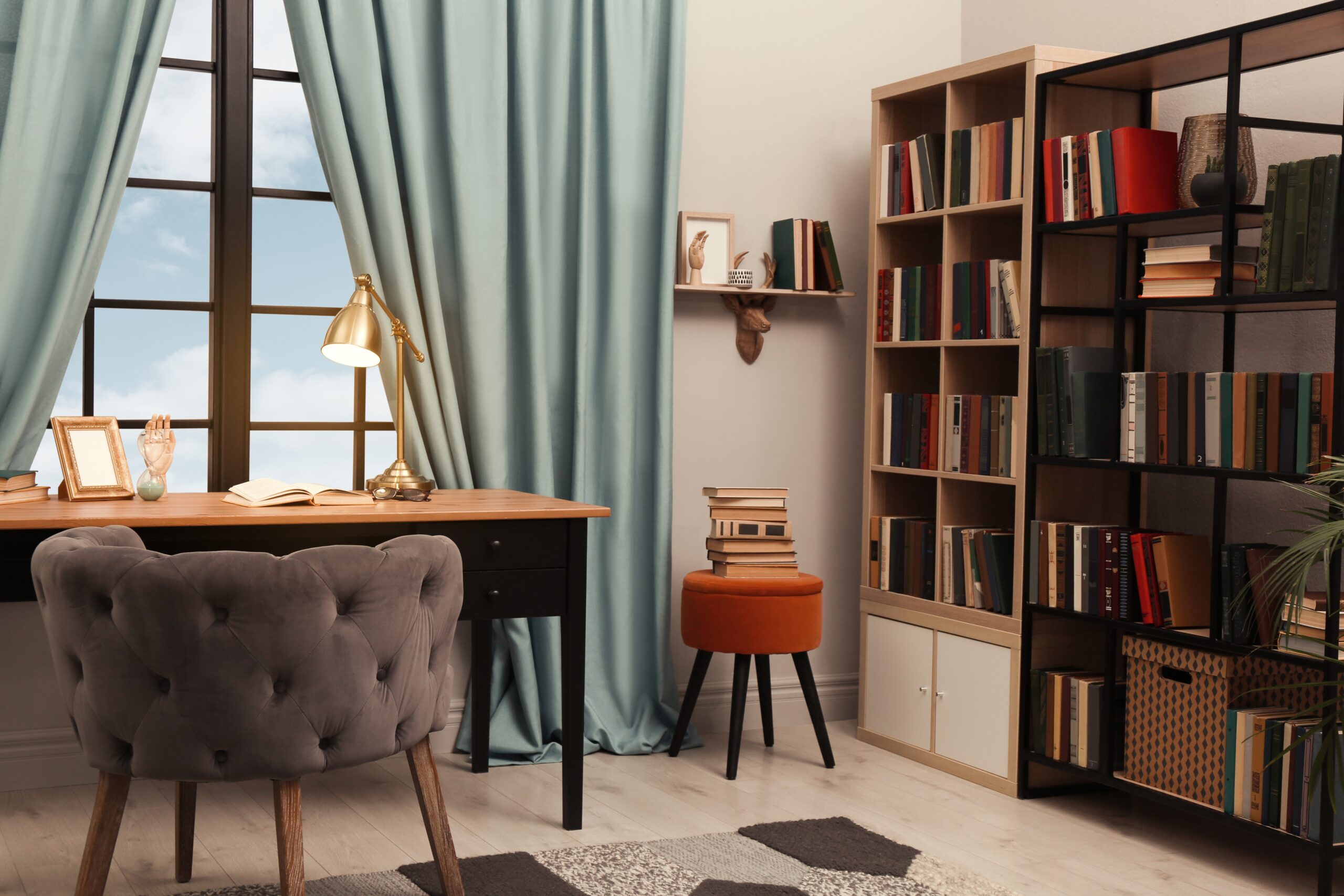 home office work environment with brown and black desk, plush tufted mauve chair, sage green curtains, modern bookshelves, orange stool with books on it, brass lamp