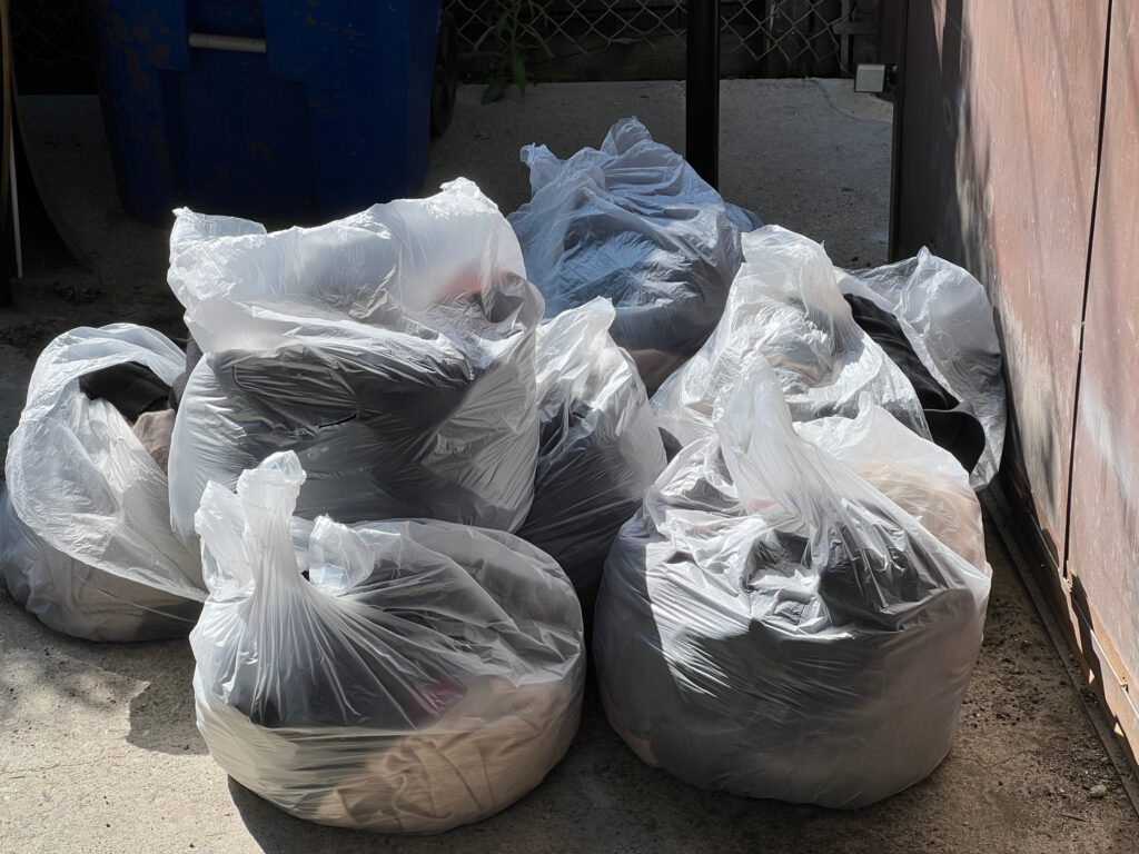 fashion brands destroy unused merchandise Clear bags of fast fashion waste from the alley by a brown metal gate Those Someday Goals