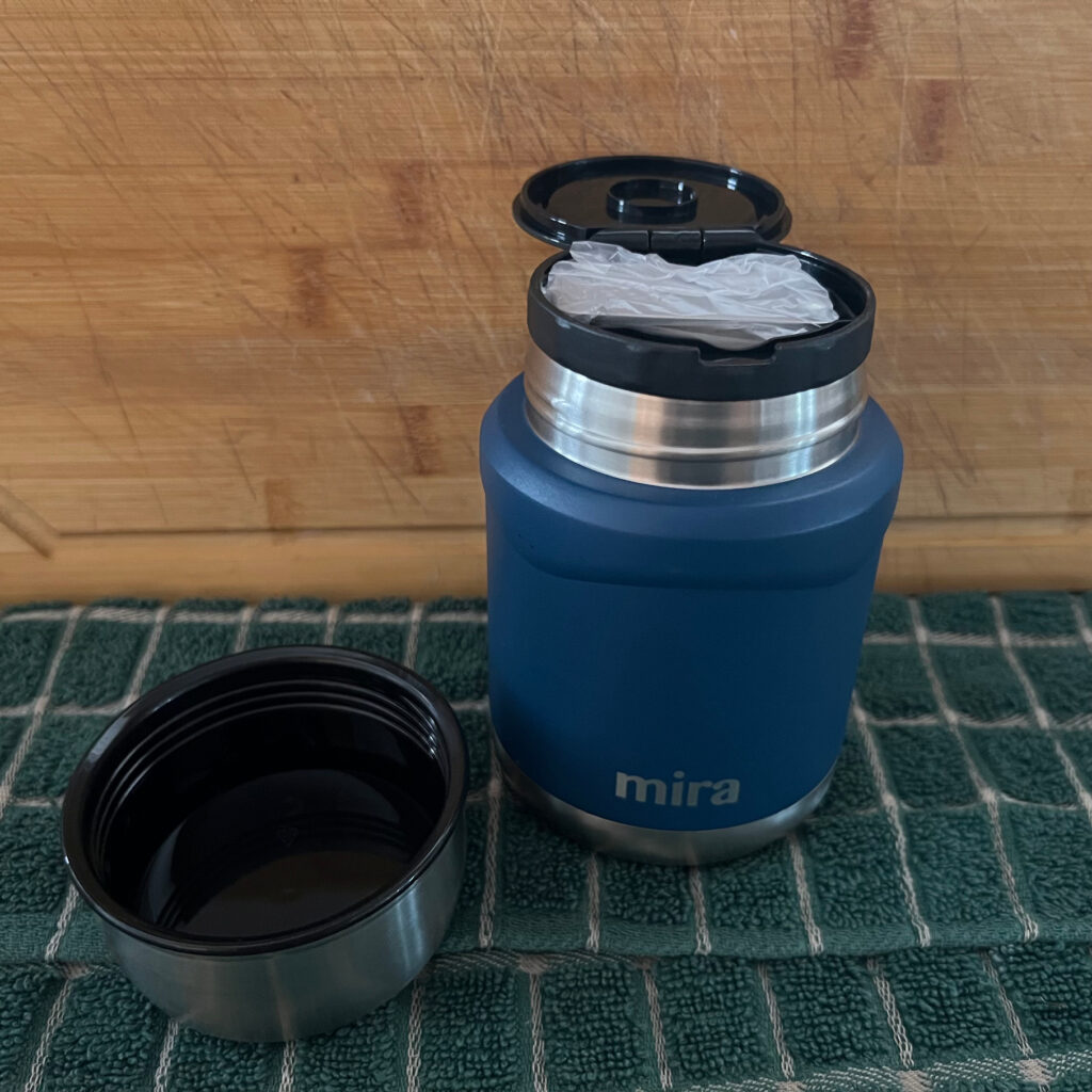 Mira insulated food jar in blue sitting on a green towel by a wooden cutting board.